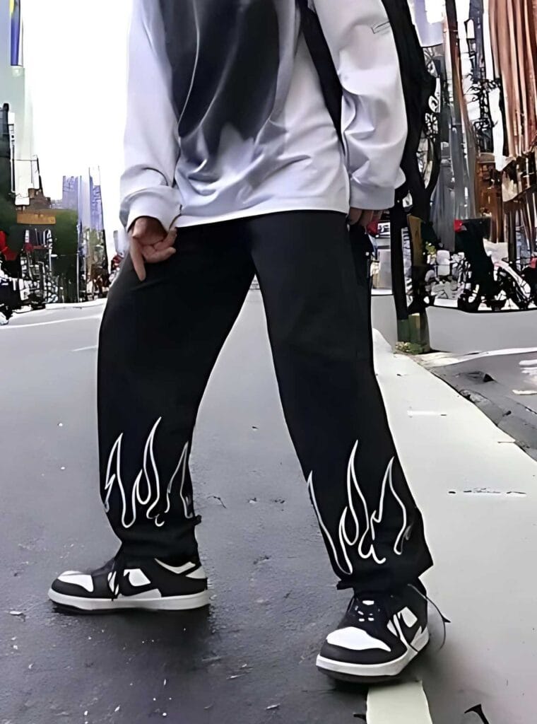 Front view of N Club men's black jeans with white flame graphics, worn with white sneakers against a grey wall